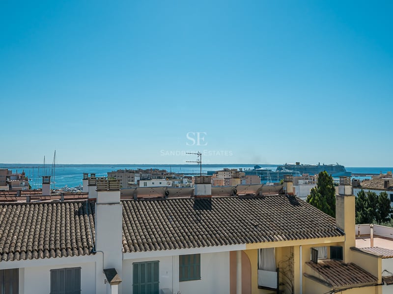 Elevated view over traditional tiled rooftops toward the blue sea with a cruise ship and sailboats in the harbor.