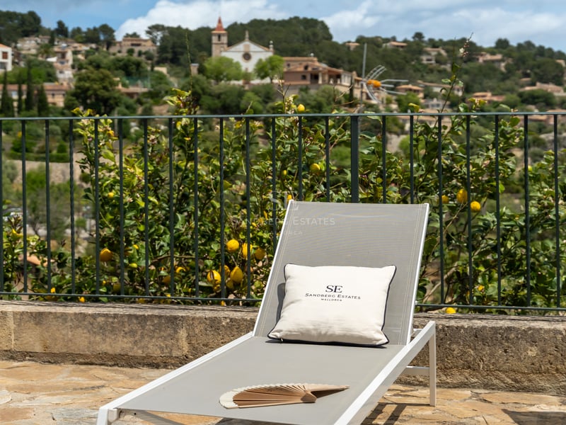 Chaise longue moderne sur une terrasse en pierre surplombant un citronnier et un village méditerranéen perché.
