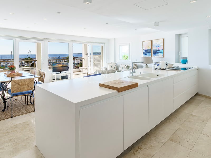 Minimalist white kitchen island leading to a dining area with a panoramic view of the harbor and sea.
