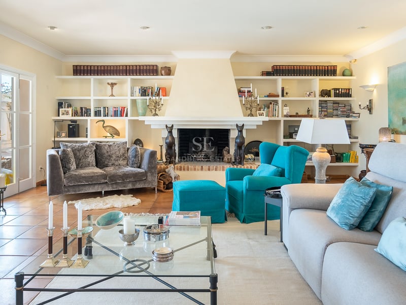 Salon lumineux avec sol en terre cuite, canapés gris, fauteuil turquoise, cheminée et étagères intégrées.