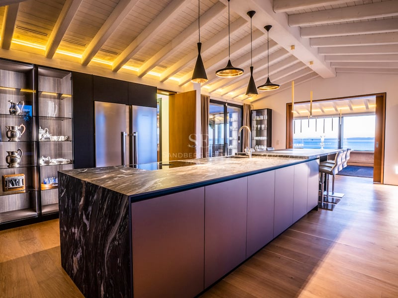 Moderne Küche mit großer Marmorinsel, Holzbalkendecke und Blick auf das Meer durch die Fensterfront.
