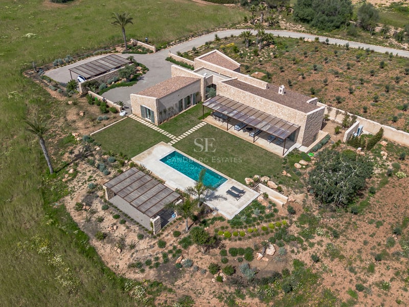 Luftaufnahme einer modernen Steinvilla mit türkisem Pool, grünem Rasen und umgebender Naturlandschaft auf Mallorca.