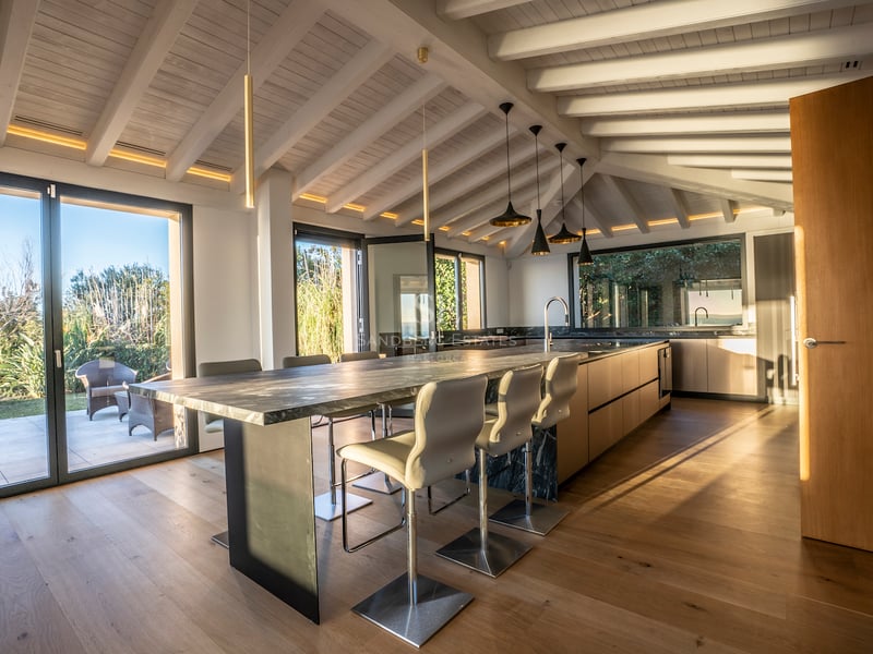 Moderne Küche mit Marmorinsel, weißen Holzbalken und bodentiefen Glasfronten zum Garten.