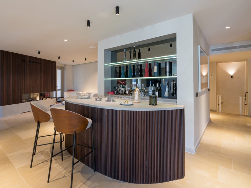 Bar incurvé en bois avec comptoir en pierre, étagères en verre éclairées et tabourets modernes.