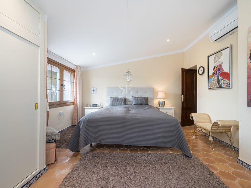 Chambre avec sol en terre cuite, grand lit avec couvre-lit gris, placards intégrés blancs et fenêtre en bois.