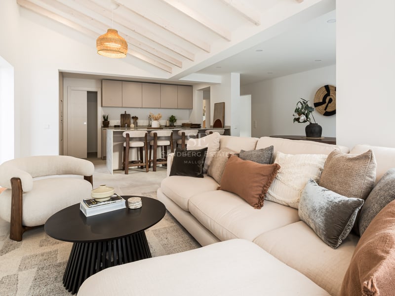 Salon lumineux avec canapé beige, poutres en bois et îlot de cuisine au second plan.