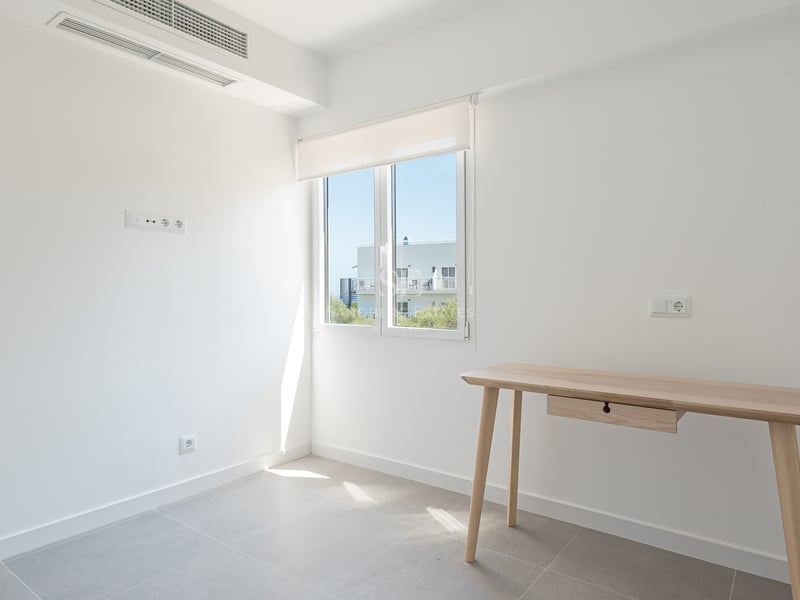 Pièce blanche avec bureau en bois clair, sol en carrelage gris et fenêtre offrant une lumière vive.