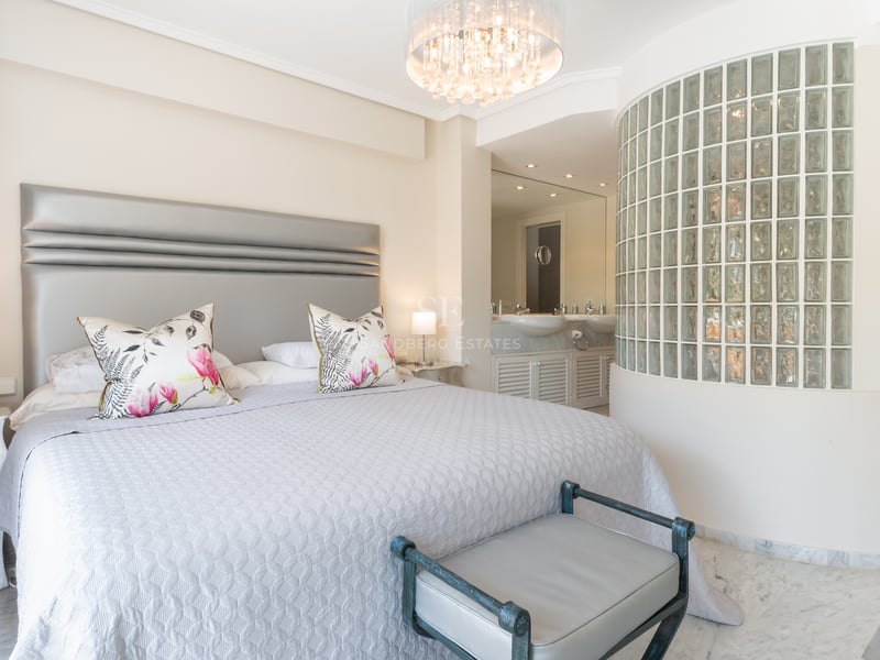 Luxury bedroom with a king bed, marble floors, a crystal chandelier, and a curved glass block wall leading to an en-suite.