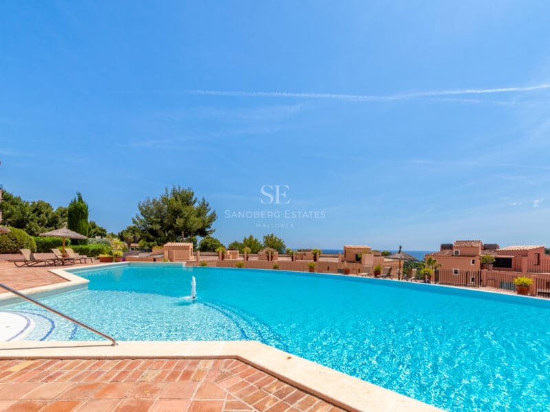 Grande piscine bleue incurvée avec fontaine, terrasse en terre cuite, parasols en paille et vue sur mer.
