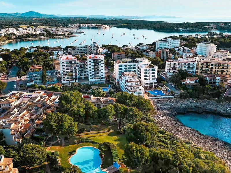 Vue aérienne de bâtiments blancs en bord de mer avec piscines, pins et une crique aux eaux cristallines.