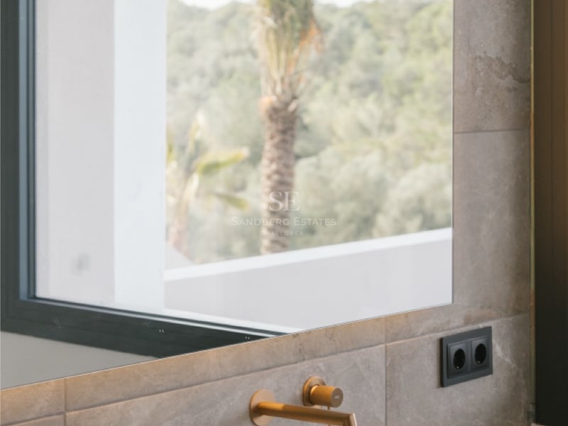 Nahaufnahme eines integrierten Natursteinwaschbeckens mit Messingarmatur und Spiegel mit Palmenreflexion.