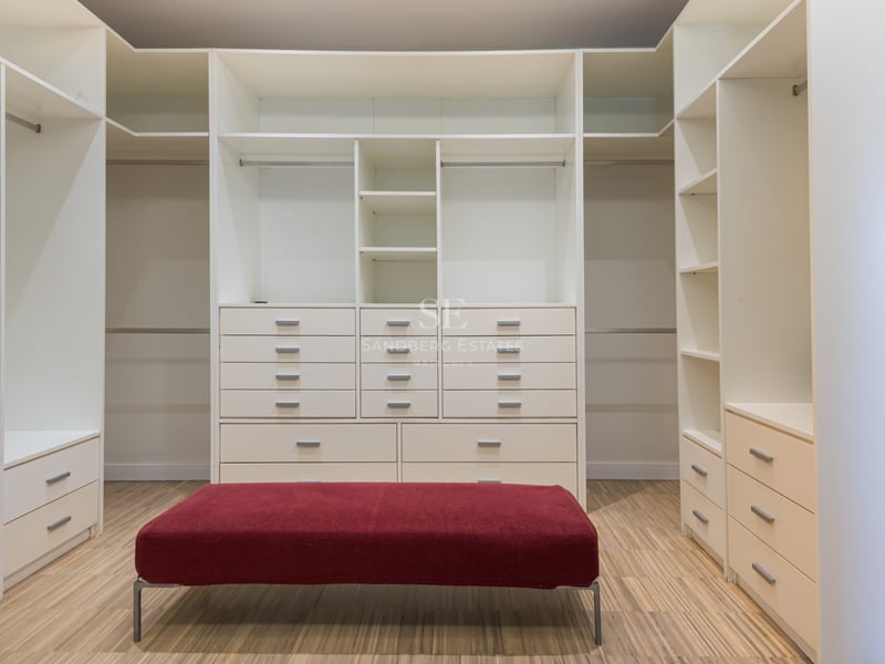 Rymlig vit walk-in-closet med hyllor, lådor, trägolv och en central röd sammetspuff.