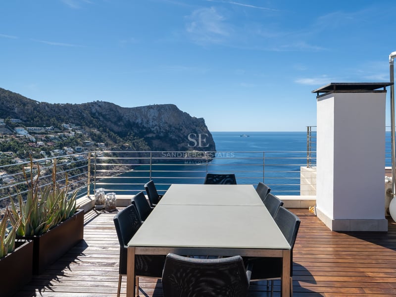 Modern rooftop terrace with a large dining table overlooking the sea and rugged coastline under a clear blue sky.
