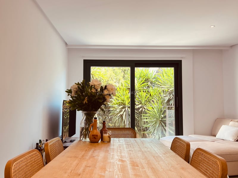 Table à manger en bois clair et chaises en rotin devant une grande baie vitrée donnant sur de la verdure.
