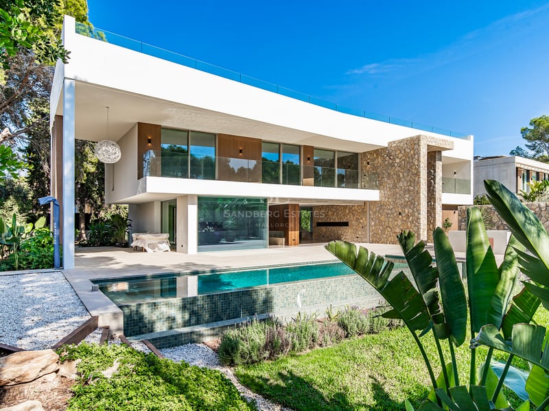 Villa moderne blanche à deux étages avec murs en pierre, piscine et garde-corps en verre par temps ensoleillé.