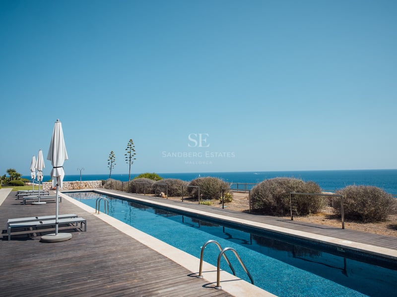 Longue piscine rectangulaire avec terrasse en bois sombre, parasols blancs et vue imprenable sur la mer azur.