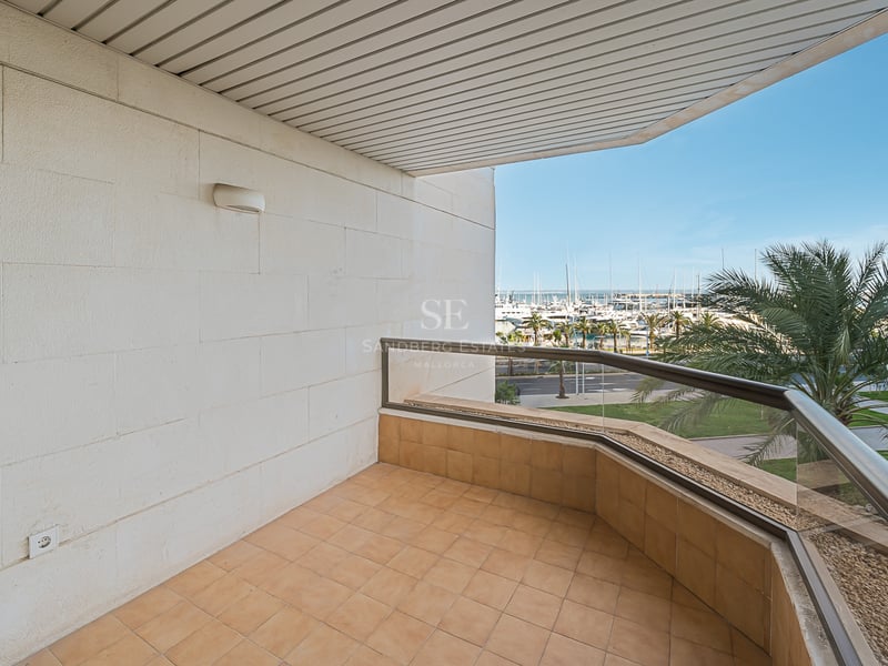 Täckt terrass med glasräcke och utsikt över en hamn med yachter och palmer under en klarblå himmel.