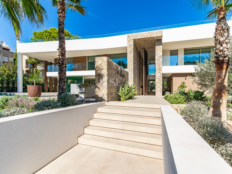 Entrée de villa blanche avec murs en pierre, escaliers extérieurs et palmiers sous un ciel bleu.