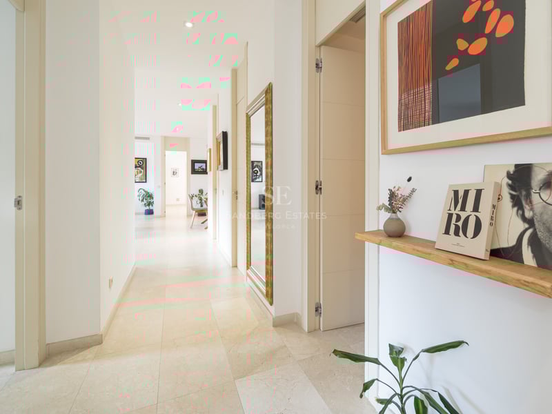 Lång vit korridor med stengolv, en stor guldinramad spegel och färgglada konstverk.