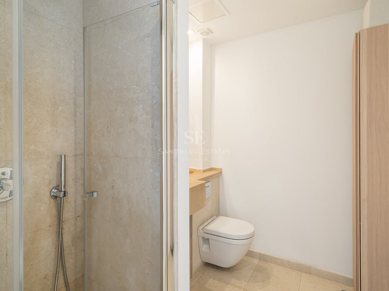 Badrum med glasdusch, vägghängd toalett och beiga stenväggar.