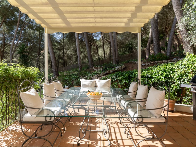 Covered terrace with wrought iron dining table, glass top, and cream cushions, surrounded by lush pine trees.