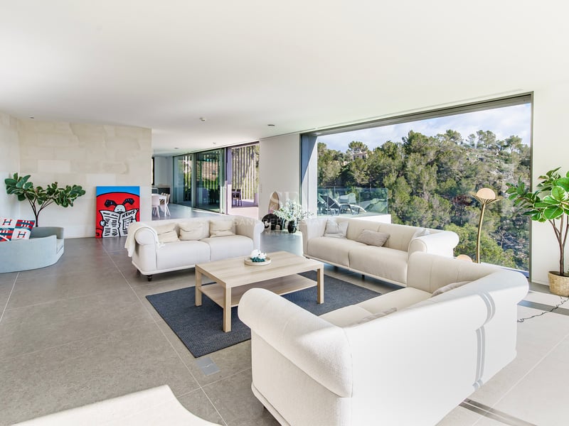 Grand salon moderne avec canapés crème et immenses baies vitrées donnant sur une forêt verdoyante.