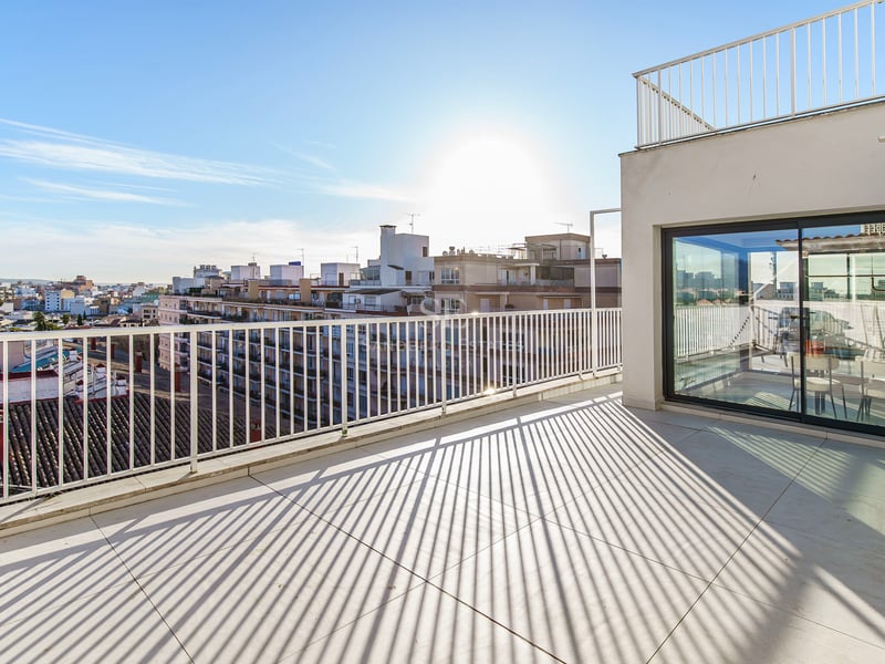 Vaste terrasse de penthouse avec carrelage et garde-corps blancs, offrant une vue sur la ville sous un soleil éclatant.