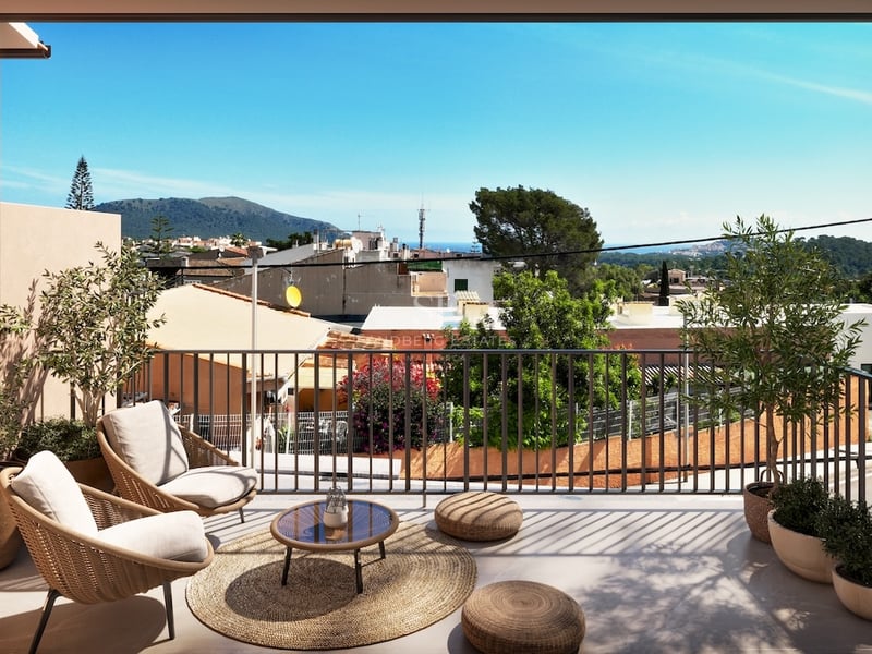 Sun-drenched terrace with wicker armchairs, olive trees, and views of a Mediterranean town and mountains.