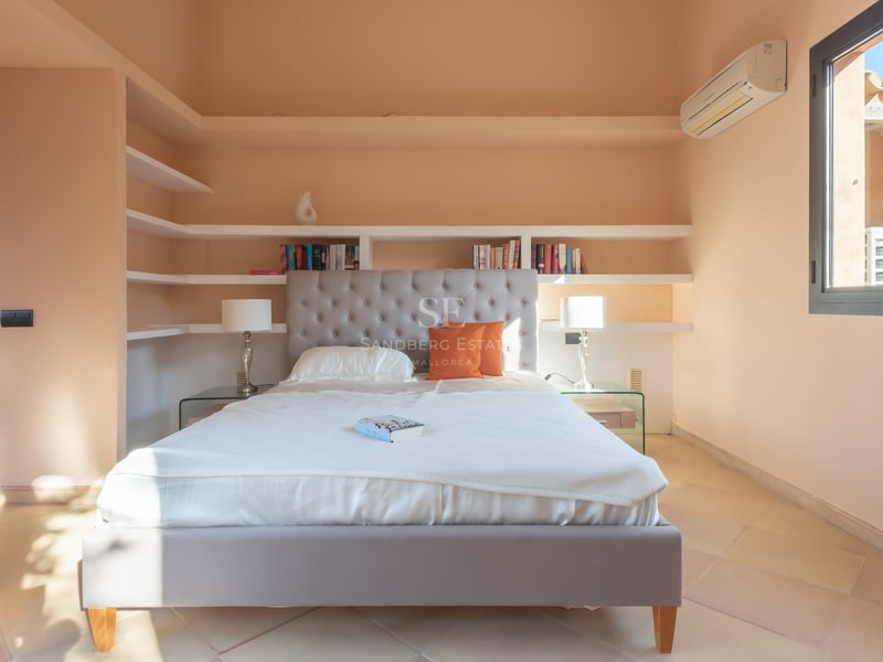 Ljust sovrum med grå tuftad sänggavel, vita sängkläder, inbyggda hyllor och terrakottagolv.