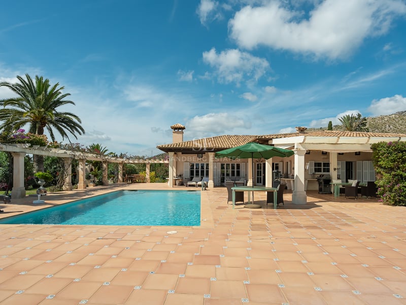 Grande piscine rectangulaire entourée d'une terrasse en terre cuite et d'une villa blanche sous un ciel bleu.