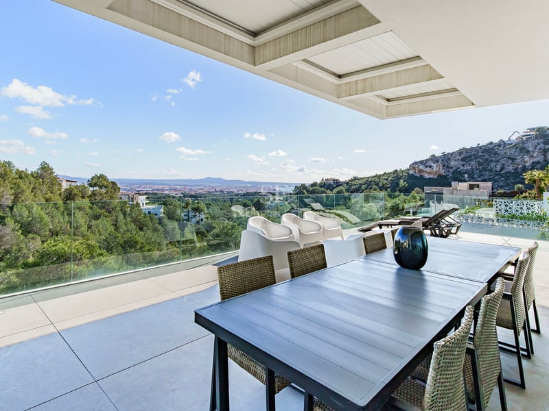 Terrasse moderne avec table à manger et garde-corps en verre surplombant une forêt et la ville au loin.