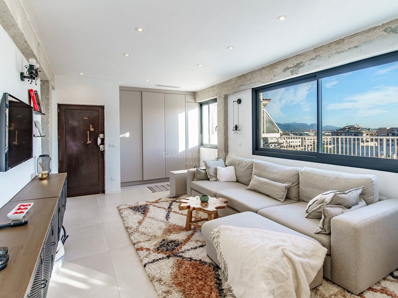 Salon lumineux avec un grand canapé gris, des poutres en béton apparent et une large fenêtre offrant une vue sur la ville.