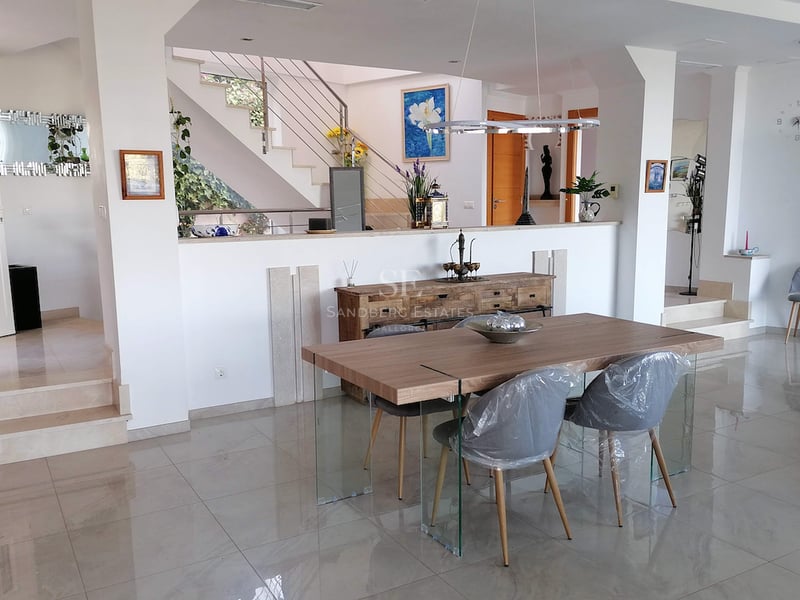 Salle à manger moderne avec table en bois, pieds en verre, sol en marbre et escaliers en arrière-plan.