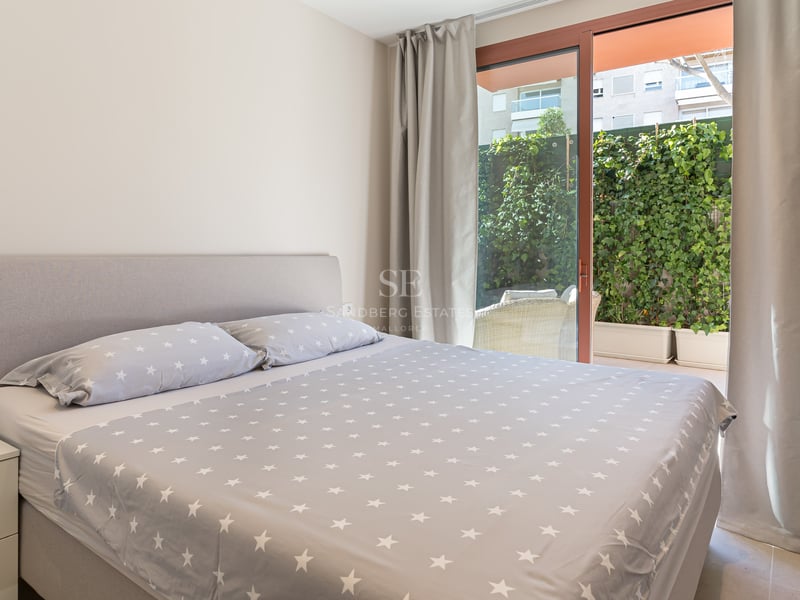 Bright bedroom featuring a grey upholstered bed and sliding glass doors opening to a terrace with lush green hedges.
