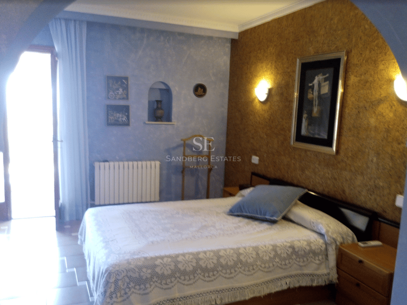 Dormitorio con colcha de encaje blanco, pared azul con textura y luz natural desde una puerta lateral.