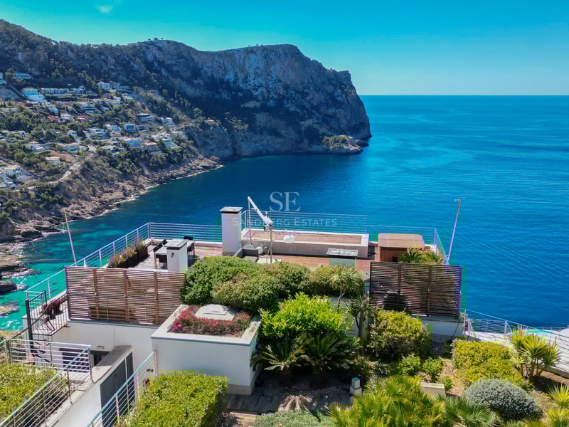 Modern rooftop terrace with glass railings overlooking a blue sea and rugged coastal cliffs.