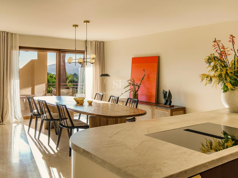 Ovaler Holzesstisch mit schwarzen Stühlen vor großen Glasfronten und einer Marmor-Kücheninsel im Vordergrund.