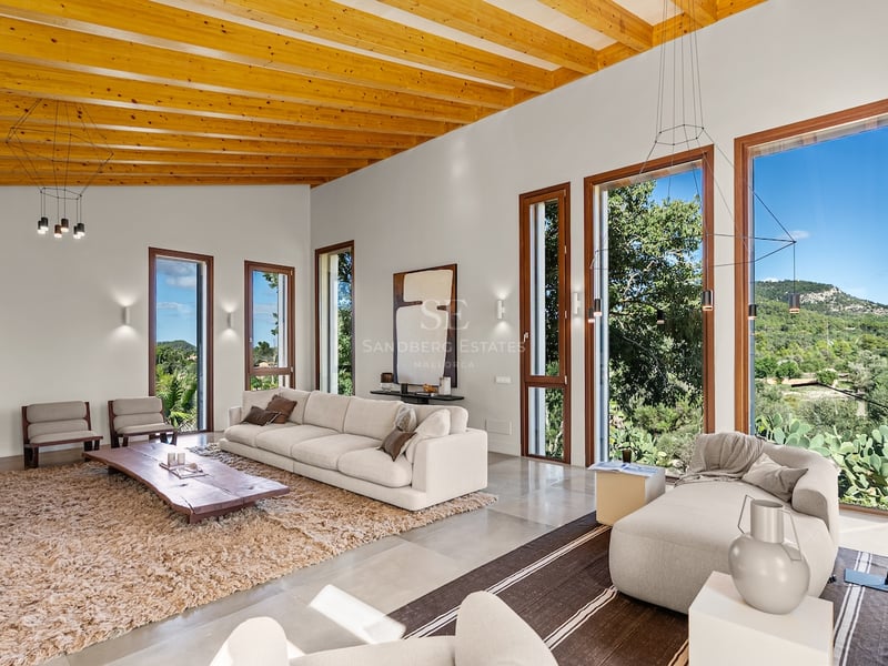 Grand salon moderne avec plafonds à poutres en bois, canapés blancs et grandes fenêtres donnant sur la montagne.