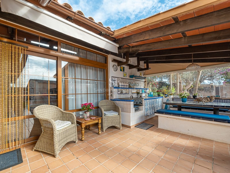 Terrasse couverte comprenant une cuisine d'été, des poutres en bois, un sol en terre cuite et des fauteuils en osier.