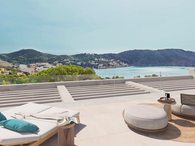 Moderne Terrasse mit Sonnenliegen und Sitzgelegenheiten mit Blick auf eine türkisfarbene Bucht und grüne Berge.