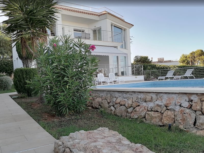 Villa blanche à deux étages avec piscine turquoise, chaises longues et mur de jardin en pierre naturelle.