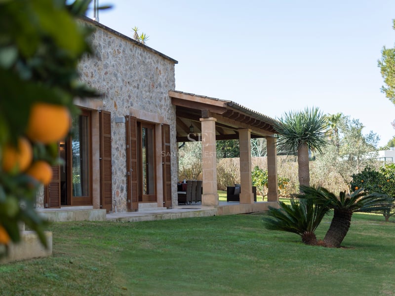 Naturstensfasad med övertäckt terrass, välskött gräsmatta och apelsinträd i dagsljus.