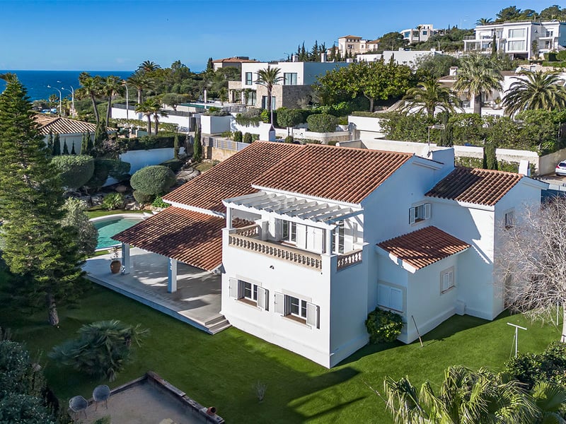 Flygvy över en vit medelhavsvilla med tegeltak, trädgård, pool och havet i bakgrunden under blå himmel.