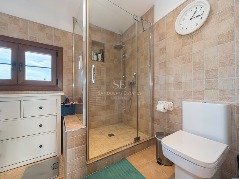 Salle de bain avec carrelage beige texturé, douche vitrée, commode blanche et fenêtre en bois.
