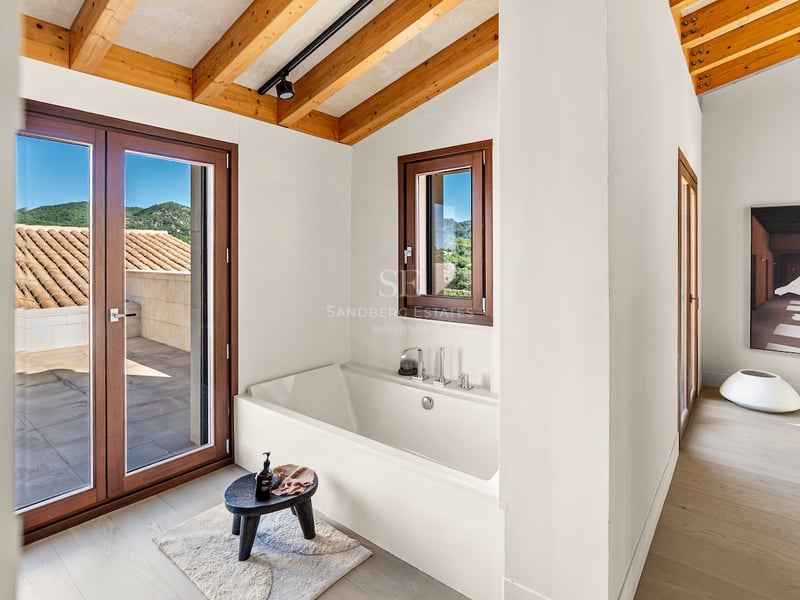 Baignoire blanche sous un plafond en poutres de bois avec accès terrasse et vue sur les montagnes.