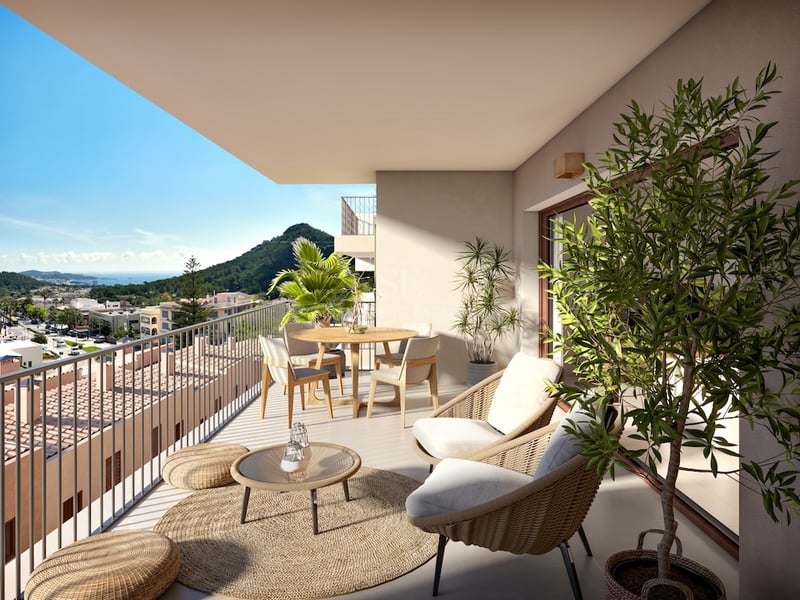 Modern balcony with woven furniture, dining table, and plants overlooking a scenic coastal town and green hills.