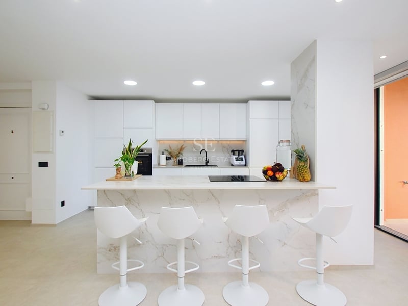 Modernt vitt kök med en stor köksö i marmor, fyra vita barstolar och inbyggda vitvaror.