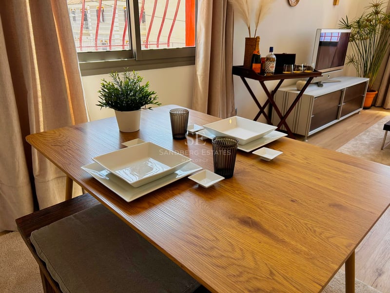 Wooden dining table set with white square plates and glasses in a bright living room.
