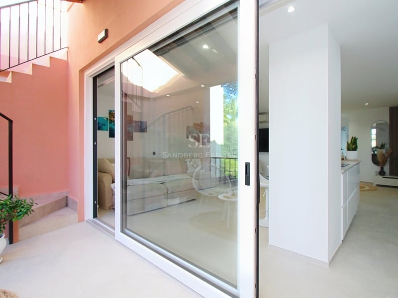 Vy från terrassen genom stora vita skjutdörrar i glas in till ett ljust modernt vardagsrum och kök.