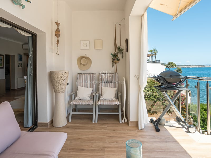 Solig övertäckt balkong med golv i träimitation, sittgrupp, grill och glasräcke med utsikt över det blå havet.